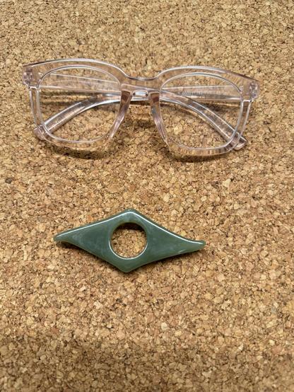 A color photo of a 3 inch long green plastic thing. It has a hole in the middle and then swooped down on either side into a triangle with a little turn at the end. It is sitting on a cork placemat and above it is a pair of pink clear reading glasses