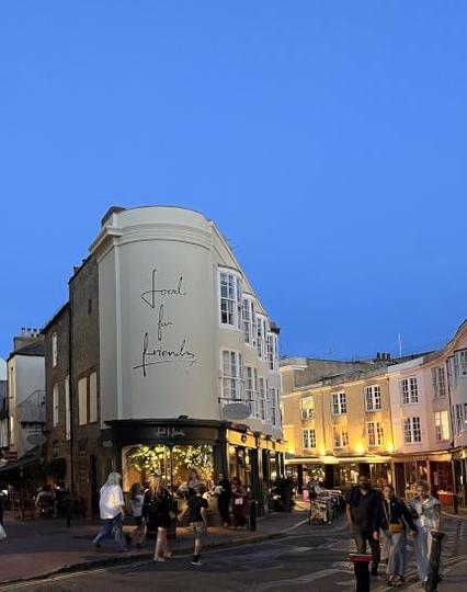 Unter wolkenlosem Abendhimmel mit letzen Blau ein altes schmales halbrundes Gebäude, unten ein beleuchteter Gastraum, die beiden Stockwerke darüber sind eine Mauer mit der Aufschrift "Food for friends", rechts davon eine schmale Gasse mit beleuchteten Altbauten.