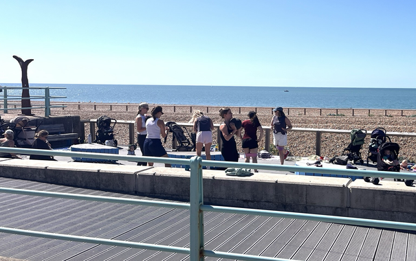 Vor sonnigem Kiesstrand und Meer eine Gruppe Frauen beim Sporttreiben, links und rechts von ihnen geparkte Kinderwagen.