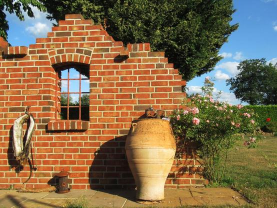 Eine Ruinenmauer aus alten Backsteinen.Sie hat ein rostiges, glasloses Stallfenster aus Gusseisen. Davor steht eine  Terrakotta-Amphore. Daneben blüht ein Strauch mit rosa Rosen, blauer Himmel.