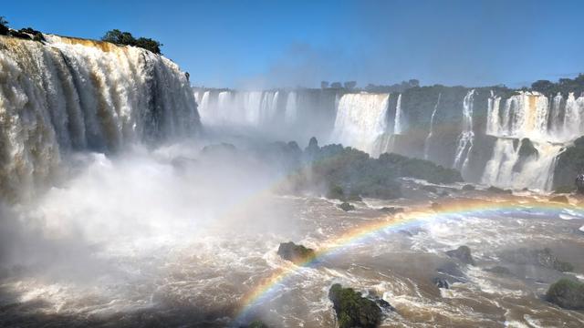 Der Betrachter steht über wild schäumendem Wasser. Im feinen Nebel darüber sind zwei Regenbögen zu erkennen. Weiter hinten stürzen gewaltige Wassermassen von einem höher gelegenen Plateau in in eine nicht einsehbare Schlucht. Blauer Himmel.