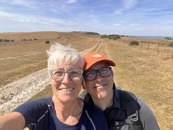 Selfie eines älteren Paars in Sonne und Wind vor kahler, gelb verdorrter Landschaft.