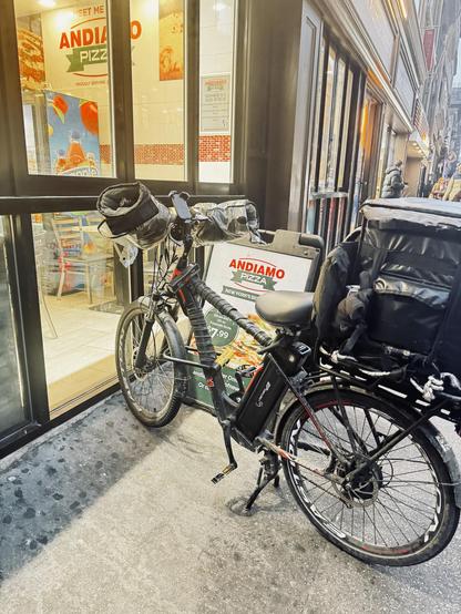 A color photo of an electric bicycle parked outside of a pizza shop on the sidewalk. The bicycle has a frame that is wrapped in black electrical tape and big gloves attached to the handlebars with plastic over them that protect your hands from the wind and cold. There is a mounted phone holder in between the handlebars and a very large insulated carrier bag on the back. It is sitting in front of a sidewalk sign that says Andiamo Pizza. It is up against the windows of the shop and again you can see the words on the Andiamo Pizza on a menu board inside.