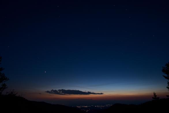 A wide angle photo of the evening twilght sky. Along the horizon there is still a band of yellow-orange sky. A small bank of clouds are a dark silhouette against the brighter sky. The sky tapers to a dark blue up high. Some stars, and one planet, are visible. Village lights are also seen. A few tree silhouettes provide more texture and interest to the scene.