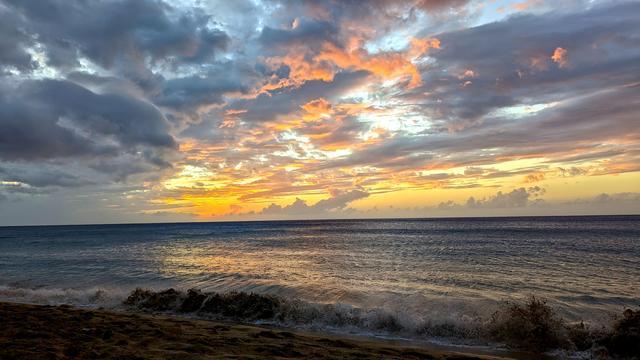 Der Betrachter steht auf einem Sandstrand und schaut aufs Meer. Im Vordergrund bricht eine Welle. Über dem Meer verschiedene kleine Wolken, die von der nicht mehr sichtbaren Sonne von unten her in orange-goldenes Licht getaucht werden.