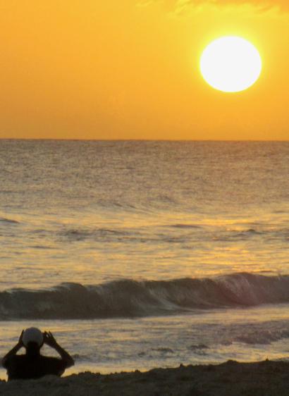 Photograph of the Sun moments before sunset over the sea horizon. 
Orange light on the sky and waves. 
In the foreground on the left, the silhouette of a bather also photographing the sunset.