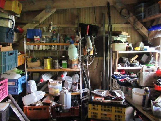Ein Schuppen mit Regalen. Alle stehen mit Kisten voll. Weitere stehen im Raum. Kanister, Dosen, Werkzeug, – ein ziemliches Durcheinander.