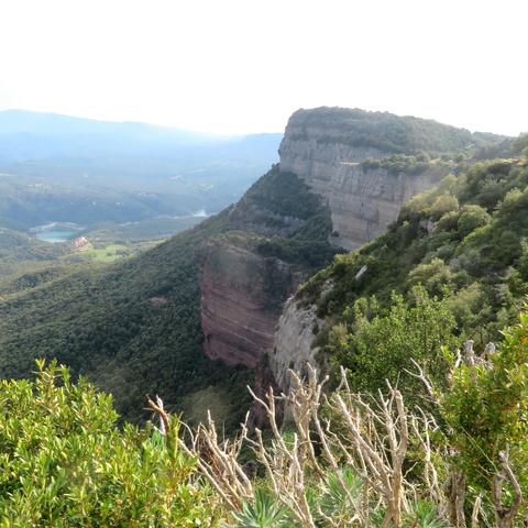 The photograph shows a section of the spectacular Tavertet cliffs as seen from above. In the background, below and in front of distant mountain ranges, is a small part of the Sant Romà de Sau reservoir.