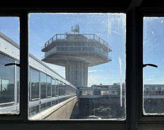 Looking through a slightly manky old window alongside a covered motorway footbridge towards the brutalist hexagonal tower at Lancaster (Forton) Services on the M6.