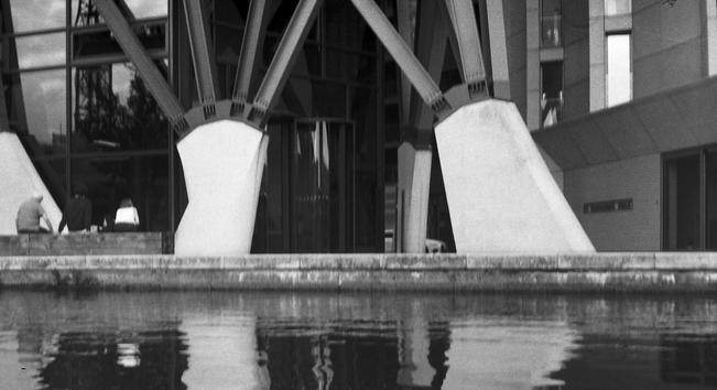 a view across a canal in london.  On the far side are huge concrete footings supporting steel pillars of a tower block.  Off to one side and dwarfed by these pillars are three people sitting on a bench, the people are looking not out across at the canal but rather at the glass of the tower block.  The canal water is reflecting some of this image. 

Camera: Fuji Auto-70 DX 
Lens: Fujinon f3.5/35mm 
Film: Agfa APX-400
Developer: 510 Pyro (1+100 for 11mins @ 20c)