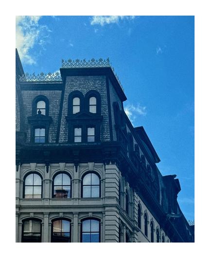 A color photo that is almost cut off diagonally from the upper Left-hand side to the lower right hand side. The upper half is a bright dark blue sky with a few clouds on the left.  The bottom part is a close-up view of the upper part of a historic building featuring ornate architectural details the top is smaller and tilts inward with black trim, shingles and a decorative roof trim. Below that are two floors of white stone with large arched windows. The side of the building can be seen going down towards the lower left hand corner.