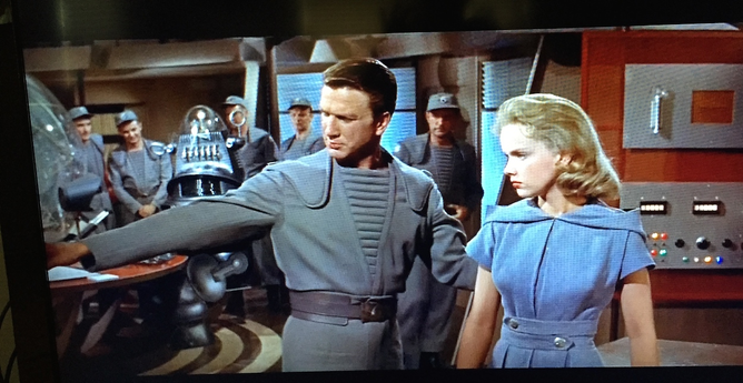 50's vintage science fiction set with Leslie Nielsen in a space corps uniform mansplaining his sleeve to Anne Francis.