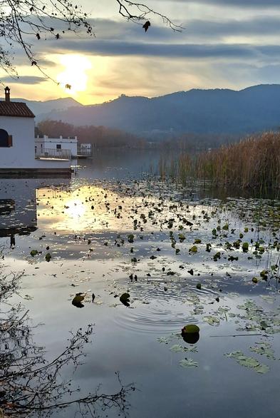 The photograph shows, against a partly cloudy sky, a sunset behind the mountains in the background, reflected in the calm waters of 'Lake of Banyoles' in the foreground. It also includes several typical lakeside structures called 'pesqueres'.