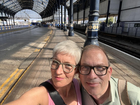 Selfie eines älteren Paars in einer Bahnhofshalle, deren Eisen-Konstruktion mit Sonnenlicht Muster auf den Boden zeichnet.