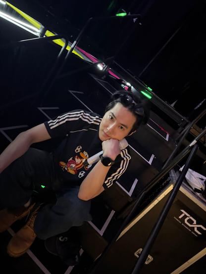 Backstage photo of kpop star SUHO sitting on black steps edged with reflective tape. He has black sunglasses on top of his head. His shirt is black with 3 white Adidas stripes down each arm and a cartoon Donald Duck with soccer balls on the front.