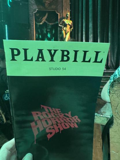 A color photo taken inside of the Roundabout Theater at Studio 54. You can see a bit of my hand holding up the play bill for the Rocky Horror Show. On the right side is the back of a seat that is wrapped in silver tape. The silver tape all over the backs of the chairs reflected some of the overhead lights casting everything in a shade of green Just above the play bill is a gold statue of a naked person with their hands on their hips (ready to do the time warp). Next to the statue is a small alcove to the side of the stage with its own curtain. That is where the narrator is during the show, played by Rachel Dratch.