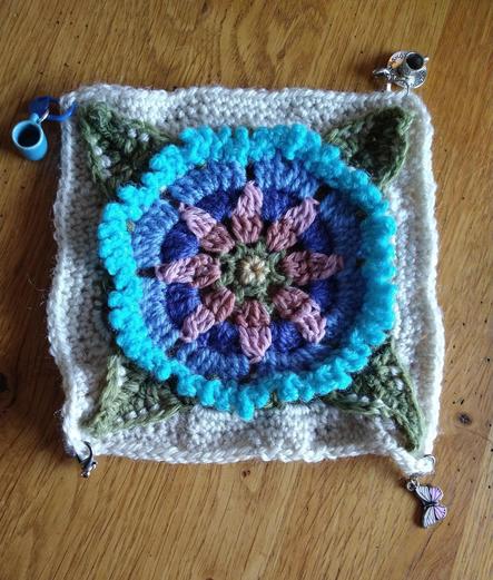 Crochet motif with a star shaped pink flower surrounded by blue petals four green leaves in the corners and a parchment coloured background