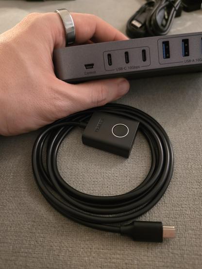 Me holding a USB switch with several USB-C and USB-A outputs, on the left edge is a mini USB connector labled "Control".
A little black box with a long mini USB cable coming out of it is placed next to it.