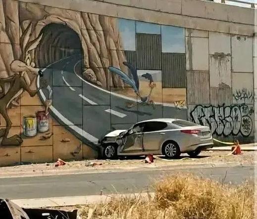 The mural features Wile E. Coyote a fake tunnel opening, directly referencing the classic cartoon gag. A silver car has crashed head on into it, crumpled, with traffic cones around it.