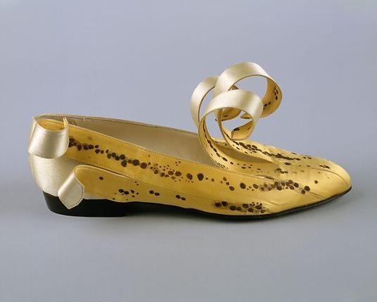 Photograph of a single yellow slip-on shoe against a neutral background; it's patterned with a very plausible series of dark brown spots arrayed in roughly linear patterns like a banana peel, and both the front (behind the toe, where the tongue would be) and heel have pieces curling off, the heel showing pale yellow silk "flesh" below.