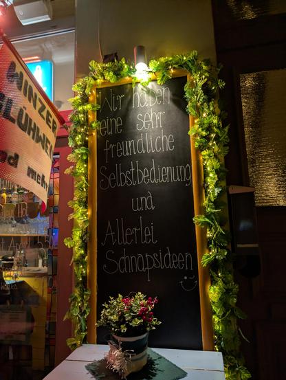 Das Bild zeigt eine an einer Hauswand angebrachte schwarze Tafel, die von einer grünen Girlande mit LED-Lämpchen eingefaßt wird. Von oben leuchtet ein Spot. Auf der Tafel steht der mit weißer Kreide geschriebene Text:
"Wir haben eine sehr freundliche Selbstbedienung und Allerlei Schnapsideen ;)"
Vor der Tafel steht ein kleiner blauer Holztisch mit einem Blumentopf mit weihnachtlicher Deko.