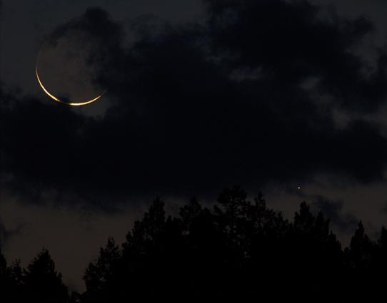 A patch of ragged low clouds take up most of the frame, but luckily the crescent moon and Mercury are visible in the faint morning twilight sky that has hints of red at the bottom of the frame. Distant conifers are a black silhouette. The sunlit crescent of the moon is overexposed and featureless but tinged yellow. Earthshine faintly shows the rest of the moon's disk.
