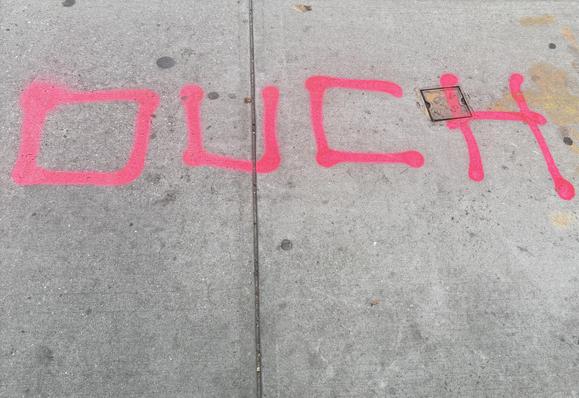 A color photo of the sidewalk and a big square orange letter somebody has written OUCH 🤕