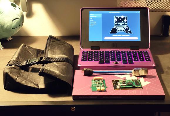Small chunky purple laptop with black ortholinear keys, a black sleeve bag, a purple print book, two small PCBs sat on the book.