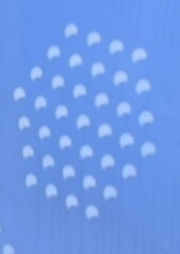 A picture of the shadow of a colander projected onto a blue card. Each hole in the colander shows the sun as a "bitten apple".  Collectively it looks like a swarm of space invaders.