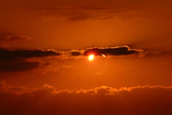 The sun is almost covered but not quite, the moon is now moving out of the way, leaving a very thin and very bright ring of light at the very top of the sun. It is illuminating a lightly clouded sky.