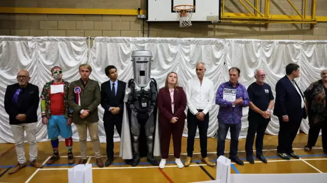 The candidates are standing in a line-up, shoulder to shoulder. Binface with his bin headgear towers above everyone else. It appears the count was held in a sports hall. There is a basketball hoop above the candidates.