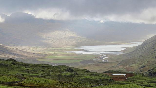 A colour photo of a landscape under a cloudy sky. The viewpoint is from height, overlooking a green valley below leading to the head of a fjord. Some areas are sunlit, showing that the cloud cover isn't complete. The foreground is a wide and rugged mossy ledge with a small tarn on the right. The distant valley below is misty with tonal perspective. The mountains on the opposite side of the valley are mostly obscured by tendrils of high cloud. It's exceptionally lovely.