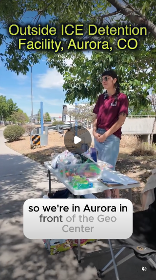 A volunteer with Casa de Paz, a pro-immigrant group in Aurora, CO. Still of the video shows a young white woman with black hair wearing a red shirt and jeans and standing behind a table full of supplies intended to help detainees' families. She's standing outside the GEO concentration camp.