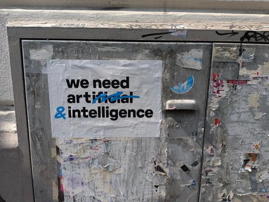 A3 großes Plakat dass auf einer Plakat- und Stickerresten übersäten Stahltür klebt. In dicken schwarzen Druckbuchstaben steht "we need artificial intelligence" - allerdings ist mit blau der hintere teil von "artificial" durchgestrichen und ein & dahinter gesetzt, womit sich "we need art & intelligence" ergibt.