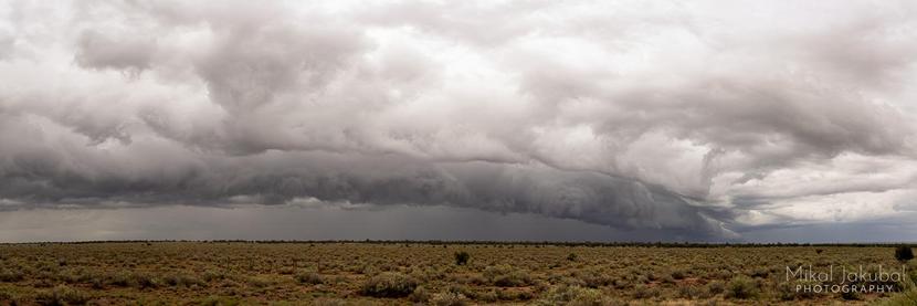 A wide panorama shot of an open plain. Above it is a broad, massive, arcing cloud, seemingly low to the ground, with a flat bottom and a shovel-like leading edge.
