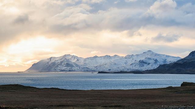 A colour photo of a seascape with snow-capped mountains in the distance. The sky is heavy with cloud but just above sea level is a gap where sunlight is streaming through, turning the clouds and the ice fields on the mountain slopes to a soft golden colour. The foreground is the shore of a fjord, rocky and with brown grasses. The expanse of water beyond is pale cyan, but is picking up a little of the colour from the sunlight. The mountain range is rugged with dark rock on the exposed edges where the wind has cleared the ice.