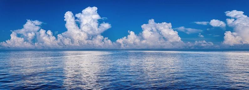A panoramic view of the ocean. At the horizon, cumulus clouds begin to build up for an afternoon thunderstorm, against a blue sky.

This photograph was created while I was on a boat, so the perspective is truly endless water and sky. The ocean is completely calm. There are no waves at all. The water is as flat as can be with only gentle ripples. The water is a deep blue, and the cloud reflections, which you can see only as smears of white, are the only thing of interest in the water.

The sky is a deep, crisp blue. Tall clouds are building toward a summer storm. There are several complexes of towers building, and the center tower is quite tall. But, for the moment, all is calm and peaceful. 

The photograph projects the vastness of the ocean, and yet, somehow, also, projects peace and calmness.