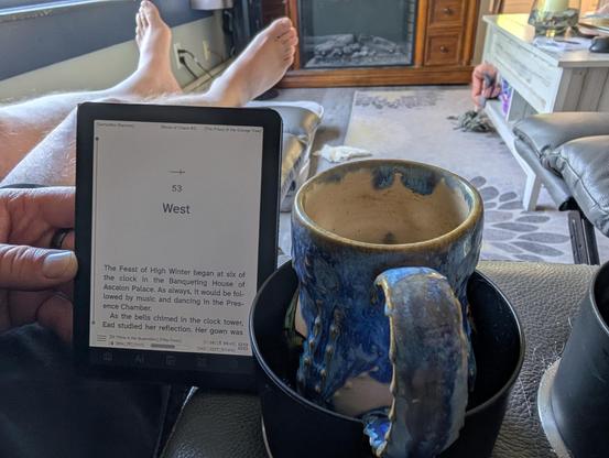 An eReader and a cup of coffee.  Some bare feet up on a recliner in the background.
