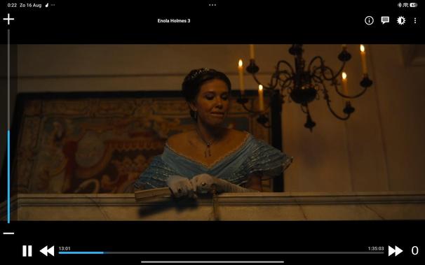 a woman watching from a balcony in dress and with candeliers suggestingin another century (actually from movie enola holms 3)