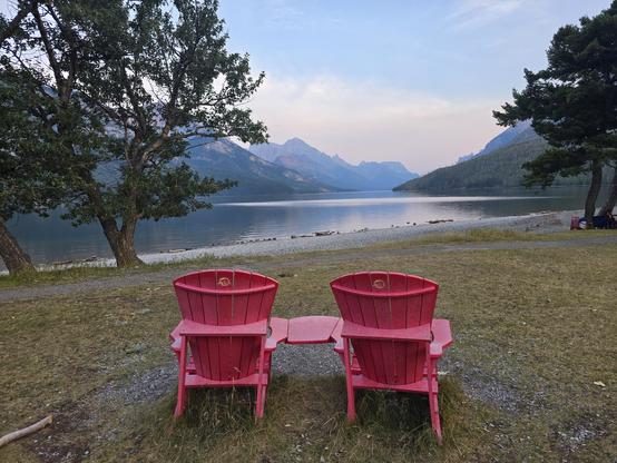 Two big red chairs facing a lake of calm water in mountains and trees.