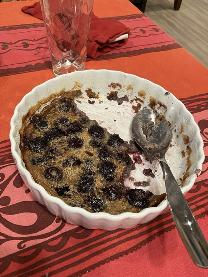 A half eaten baked good in a white dish with scalloped edges and a spoon sticking out. The food is dark golden brown and mottled with dark specks (those are the cherries). The spoon has remnants of the custard and its grey-purple.