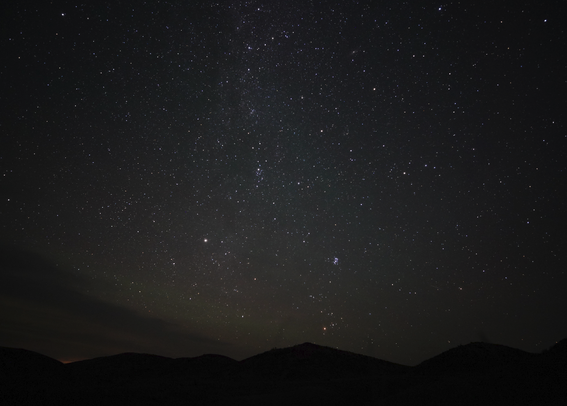 A stunningly clear picture of the stars above perfectly dark silhouetted hills.  The Milky Way is vertical coming up from the horizon in the middle of the photo, the Pleiades easily visible.