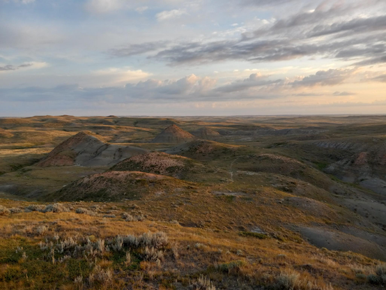 A view looking out over weird hills and badlands, perfectly lit by the rising sun, stretching off to the perfectly flat horizon under a blue sky with a few lovely fluffy clouds.