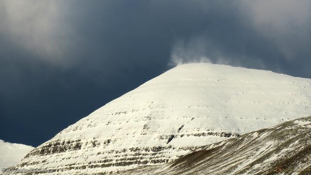 A colour photo in a monochrome palette of a snow-covered mountainside under a dark sky. Layers of rock are just visible through the ice, and parts of neighbouring peaks are also in the shot. From the pinnacle of the rounded mountain, a cloud of snow has been lifted into the air above by the buffeting winds.