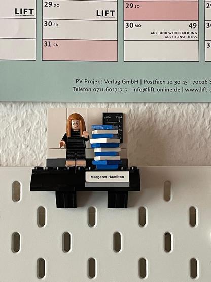 A Lego minifig of Margaret Hamilton, recreating the famous paper stack photo (from the Lego Women in NASA set), attached to an Ikea Skadis board on a white wall.