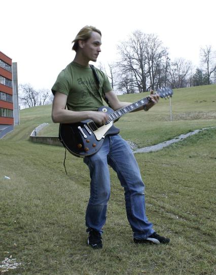 Picture of a scrawny young guy in a green t-shirt, jeans, and sneakers "playing" the guitar (without cord) on a sickly green winter lawn behind the campus of the FH Salzburg.