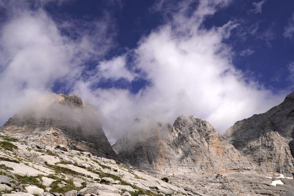 Three rocky mountain peaks (limestone) surrounded by some clouds with also some blue sky visible. We are in the alpine and it is mostly rock but there is some greenery (grasses etc) as well as a snowpatch to the right.