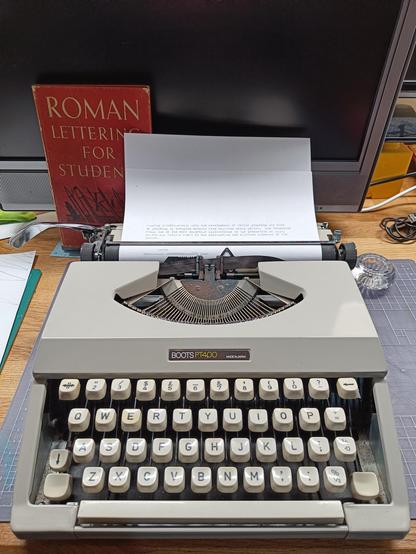 Smart compact looking typewriter on my desk. A piece of paper is in the typewriter, with a couple of sentences of text typed out. From the book "Roman Lettering for Students", which is propped up next to the typewriter.