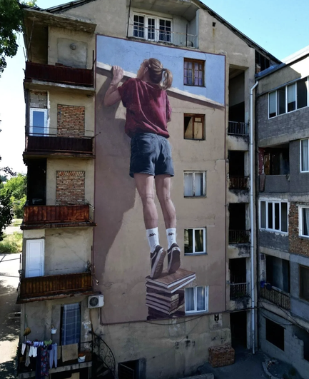Concrete Horizons mural by Edoardo Ettorre in Gurjaani, Georgia, showing a child balancing on books to peer over a painted ledge.