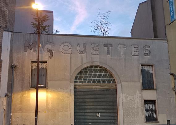 a building with a run-down sign that used to say “moquette”, the font is chunky and round, the kerning is naïve, most letters are missing but their mark is still visible, the O has a chunk of letter hanging loose